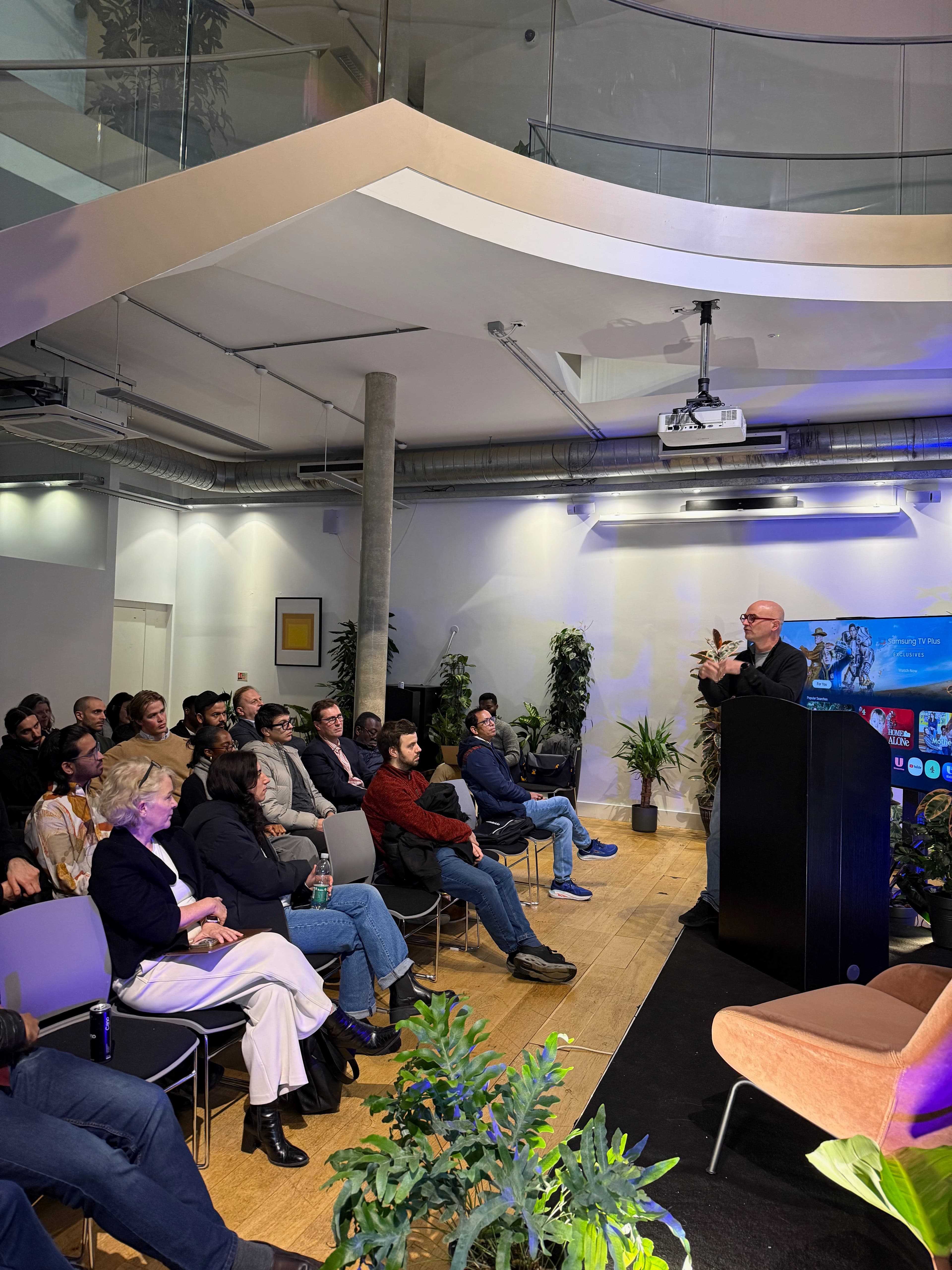 Speaker presenting to a seated audience beneath a curved staircase